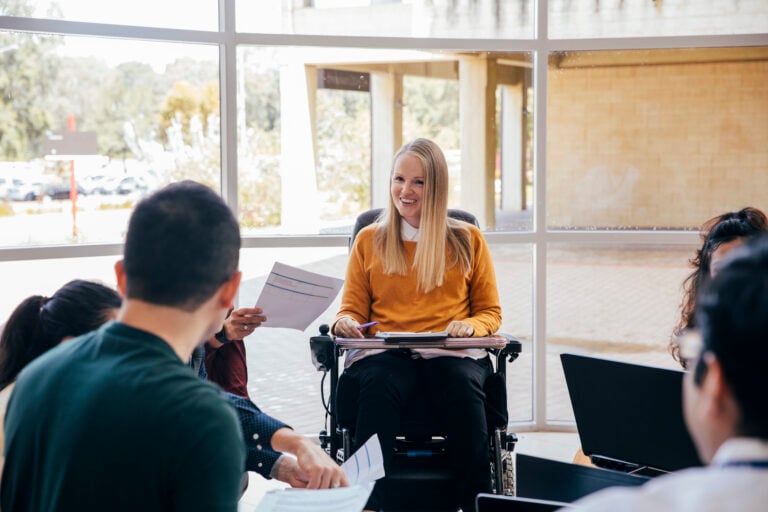 A female wheelchair user with Muscular Dystrophy can be seen smiling in a group meeting. Someone who is out of shot can be seen handing her a document.