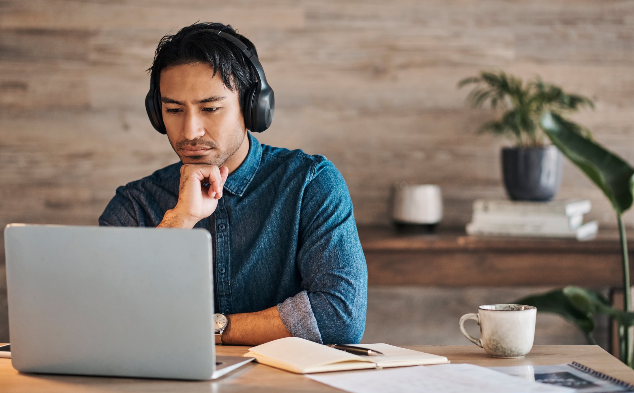 A man wearing over-ear headphones sits at a desk, focused on a laptop with his hand resting thoughtfully on his chin. A notebook, pen, and coffee mug sit on the desk, while a plant and books are visible in the softly blurred background of a home office or workspace.