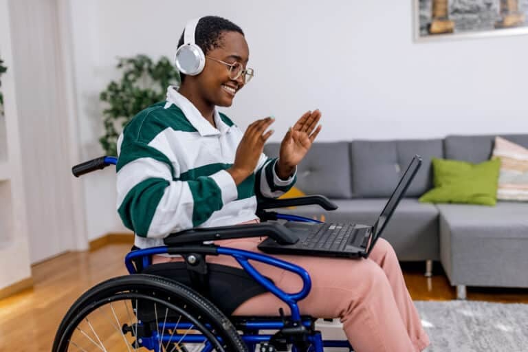 A young African American woman in a wheelchair is relaxing in her living room, with a laptop on her lap, cheerfully smiling.