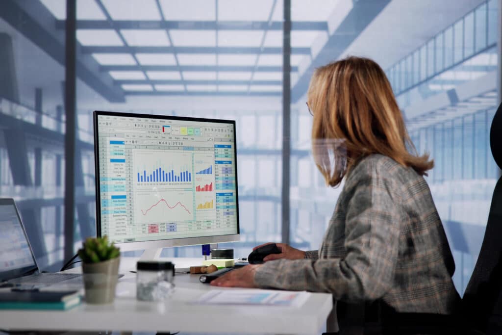 A woman sits at a desk in a modern office, viewed from behind and slightly to the side, using a desktop computer displaying spreadsheets, charts, and data visualizations. A laptop, small potted plant, and office items sit on the desk, while large windows and a bright, open workspace fill the background.