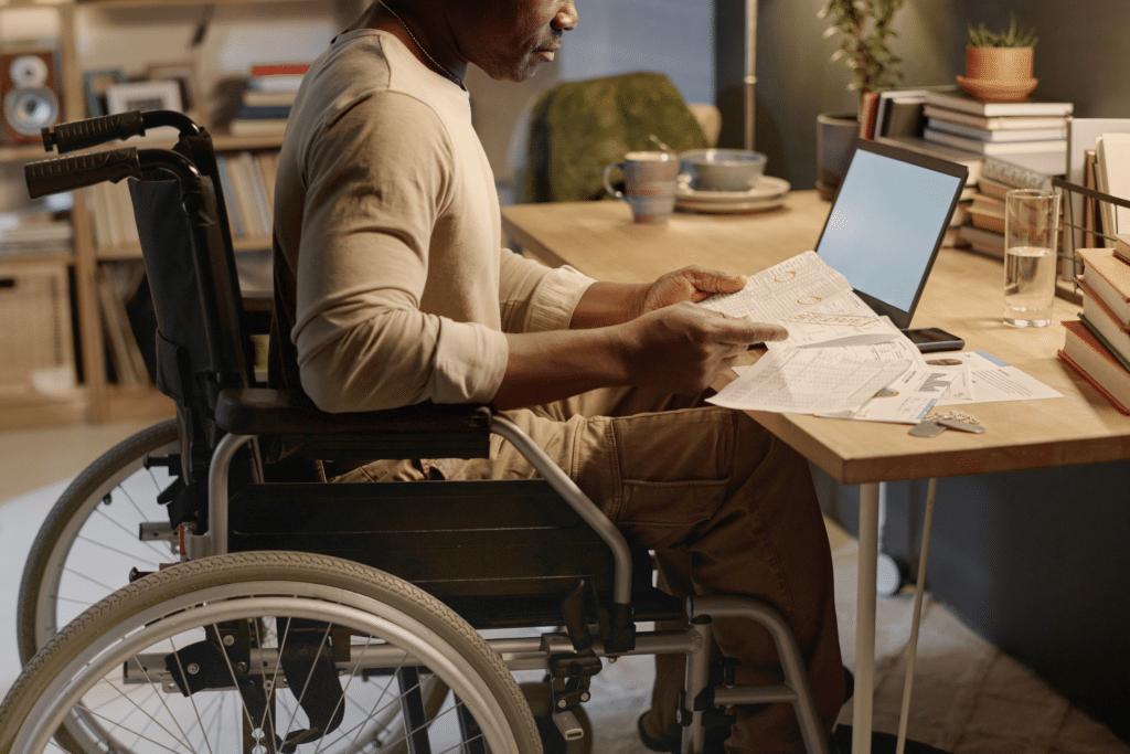A man seated in a wheelchair at a wooden desk reviews paperwork beside an open laptop, with books, a mug, and a glass of water arranged around a home office workspace.