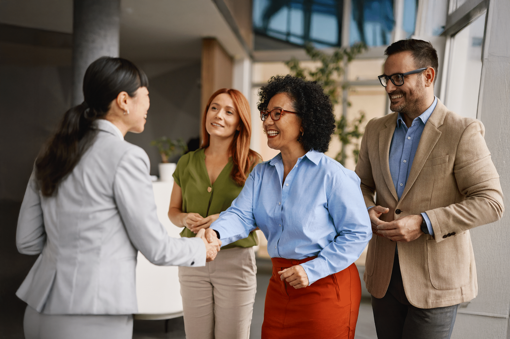 Four professionals converse in a bright office as two women shake hands, with two colleagues smiling nearby.