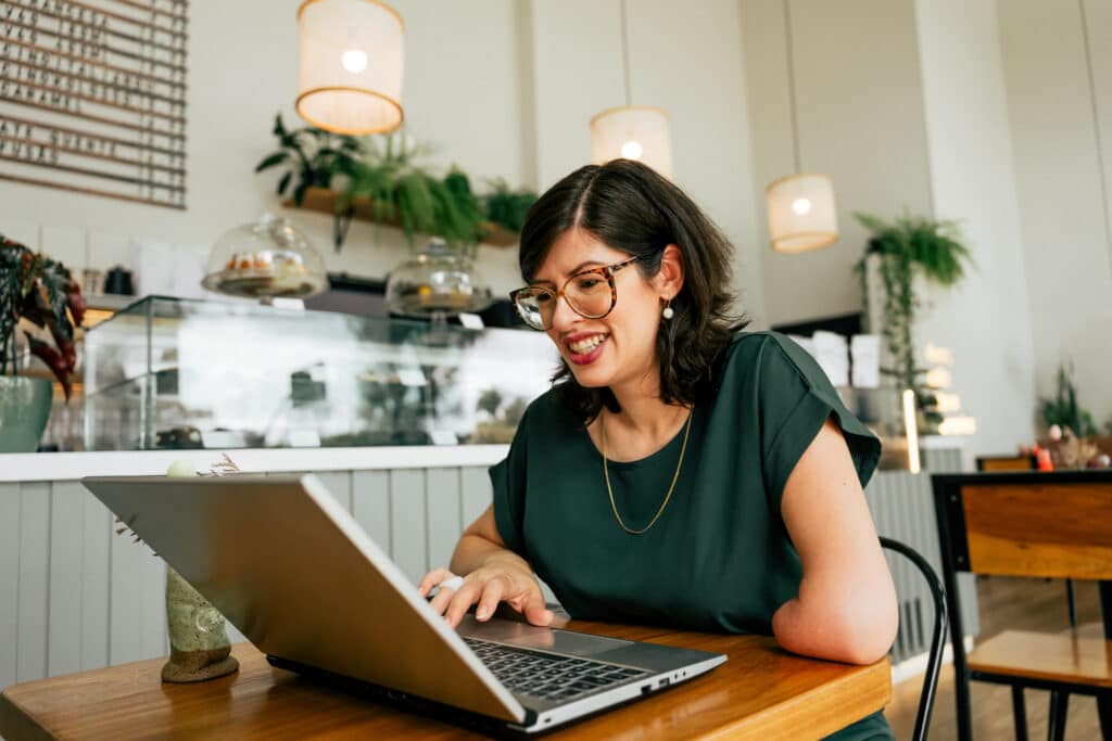 Woman with glasses and a visible upper-limb difference smiles while working on a laptop at a table in a bright café with plants and hanging lights in the background.