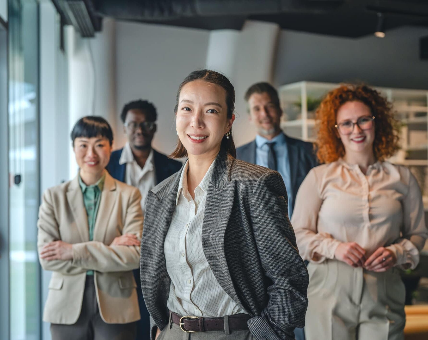 A diverse group of five professionals stands together in a modern office. A smiling woman in a gray blazer stands confidently in the foreground with her hands in her pockets, while four colleagues stand behind her, smiling toward the camera.