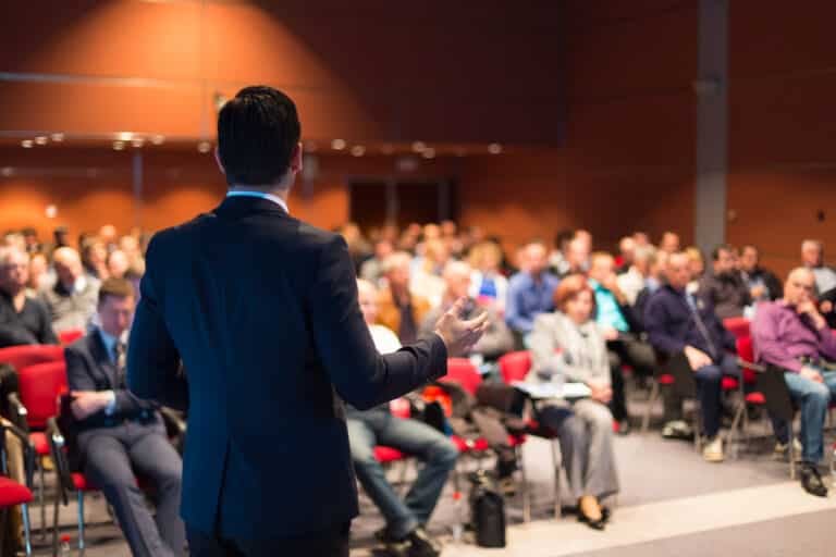 Speaker shown from behind in front of a large audience seen out of focus, inside a theater-style room.