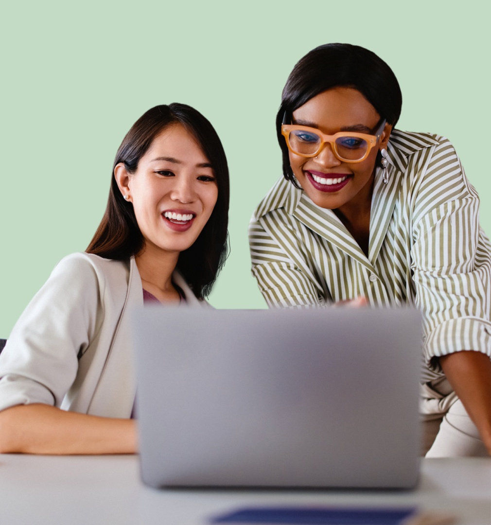 Two women smiling and looking at a laptop. One wears a light blazer, the other a striped shirt with orange glasses. The mood is friendly and collaborative.