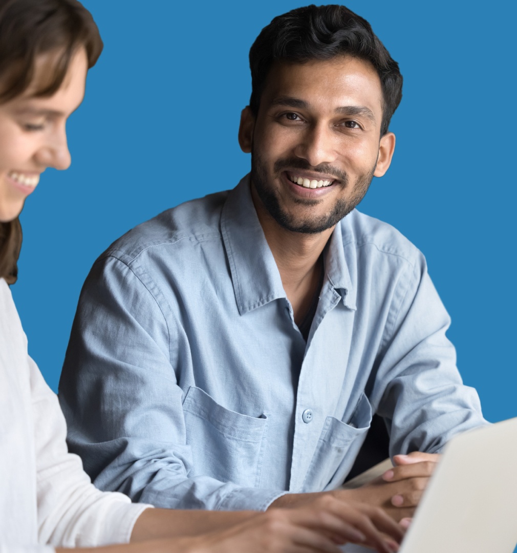 Smiling man in a light blue shirt sits at a table, looking directly at the camera. A woman beside him in white is slightly blurred. Blue background.