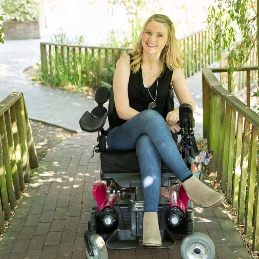 A photo of a blonde woman sitting in a wheelchair with her legs crossed, smiling.