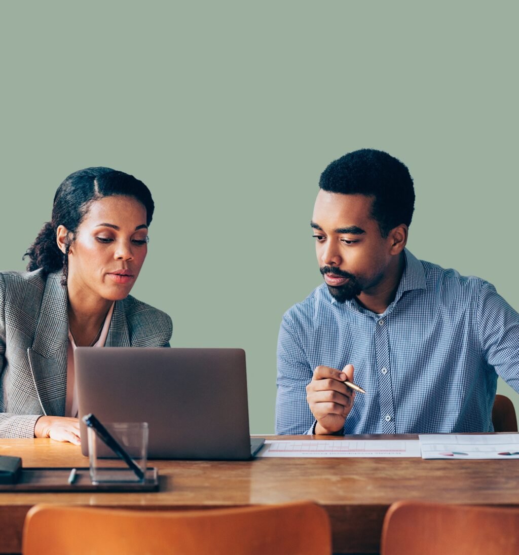 A man and a woman in business attire working together at a table, reviewing information on a laptop and analyzing printed charts.