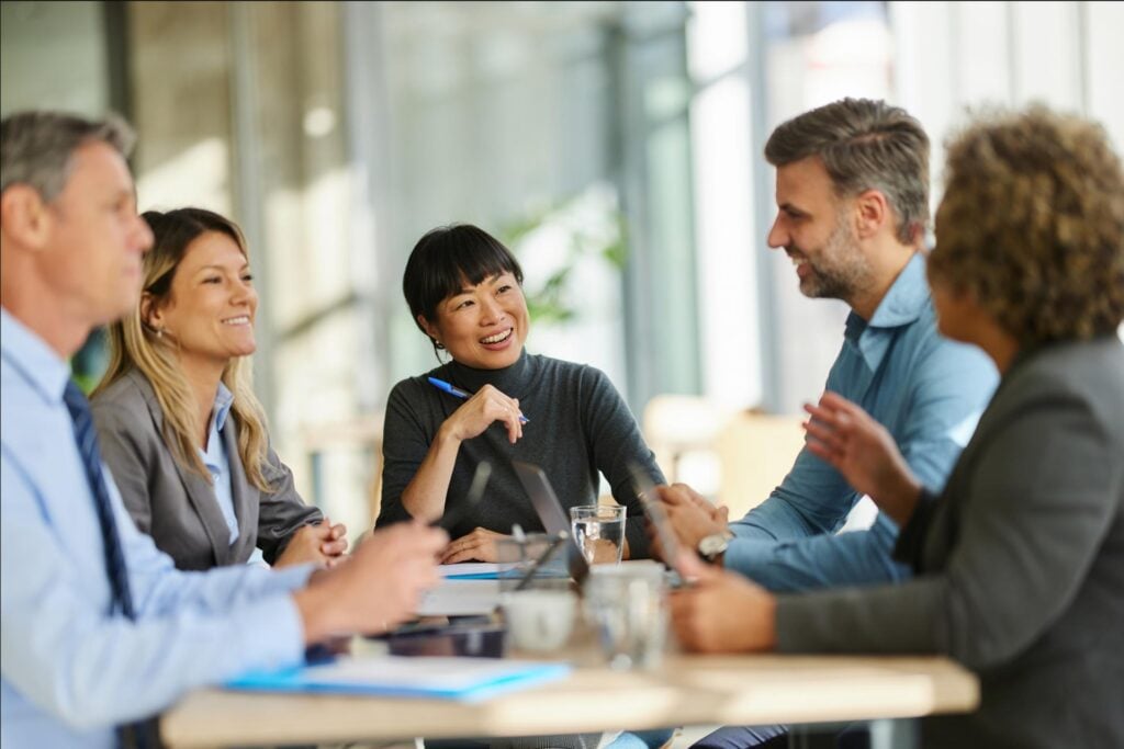Five professionals sit around a table engaged in a lively and friendly discussion. They are dressed in business attire and appear to be in a modern office setting with natural light. One woman in a dark turtleneck is smiling and holding a pen, while others listen and contribute to the conversation. Laptops, notebooks, and coffee cups are on the table.