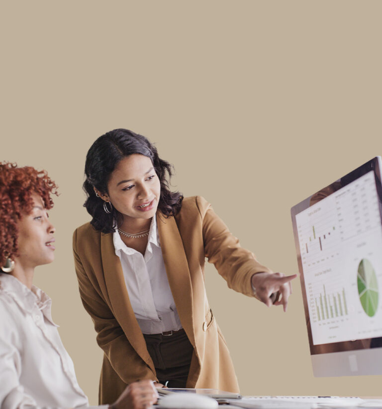 Two businesswomen collaborating at a desk, one pointing to data visualizations on a desktop monitor showing bar and pie charts.