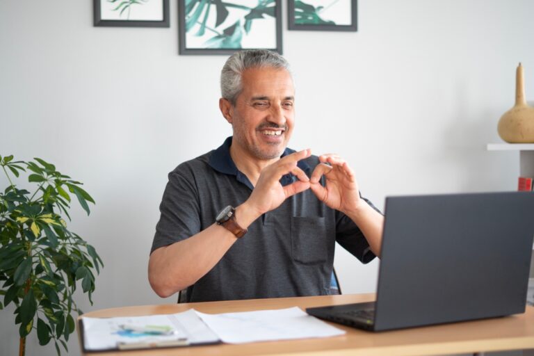 A smiling man with gray hair sits at a desk in front of a laptop, using sign language during a video call. He is in a bright, home office environment with plants and framed artwork on the wall behind him.