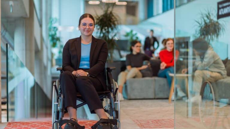 A confident woman in a wheelchair, wearing a suit and glasses, smiles in an office with colleagues chatting in the background. The atmosphere is inclusive and professional.