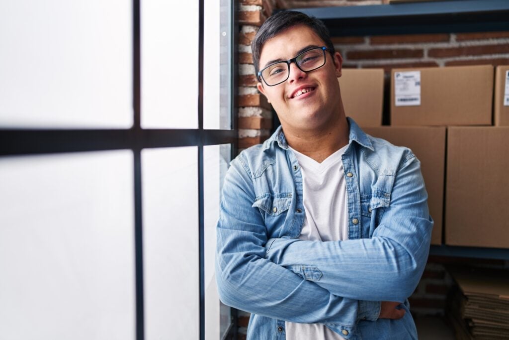 A smiling young man with glasses and dark hair stands with his arms crossed, wearing a light blue denim shirt over a white t-shirt. He's positioned in front of a large window with black frames and exposed brick walls, with cardboard boxes stacked on shelves visible in the background, suggesting a warehouse or storage facility setting.