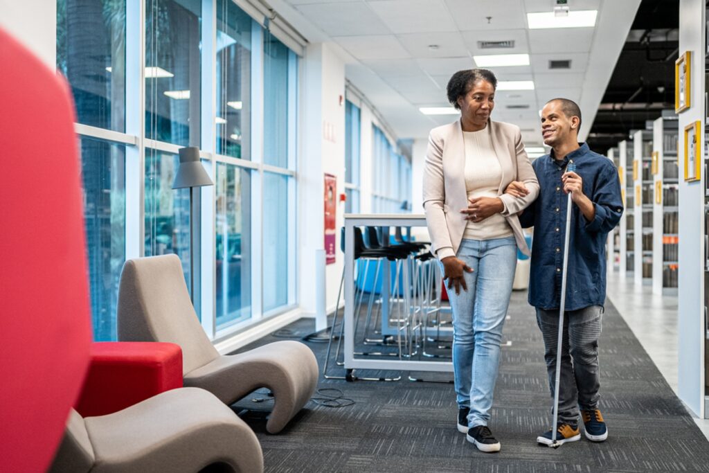 A woman in a beige blazer and jeans walks alongside a man using a white cane in a modern office hallway. The space features large windows, contemporary seating with red and beige chairs, and a polished floor. The man is wearing a dark blue shirt and appears to be having a friendly conversation with his companion as they move through the bright, well-lit corridor.