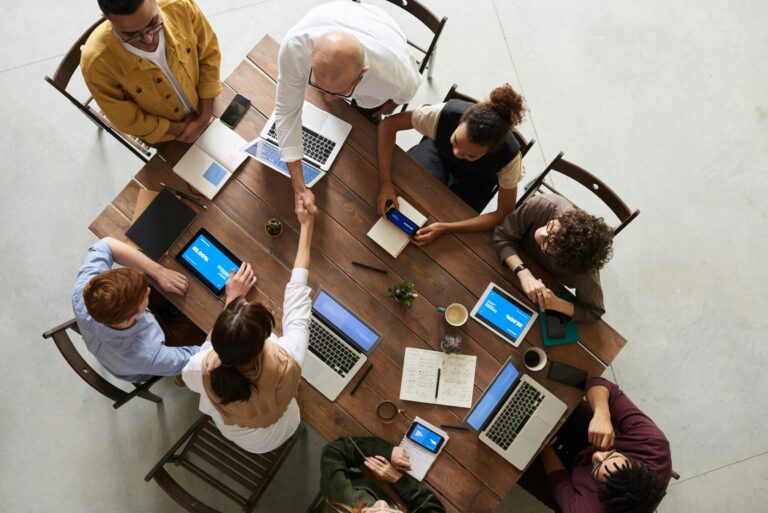 An aerial photo of eight people sitting around a table. Two are standing, shaking hands and the rest appear engaged. There are many laptops, devices, and notebooks open on the table.