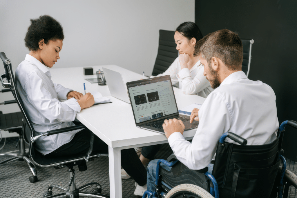 Three colleagues sit around a conference table working on laptops and taking notes during a meeting. One man uses a wheelchair and works on a laptop while two coworkers review documents and write in notebooks in a modern office setting.