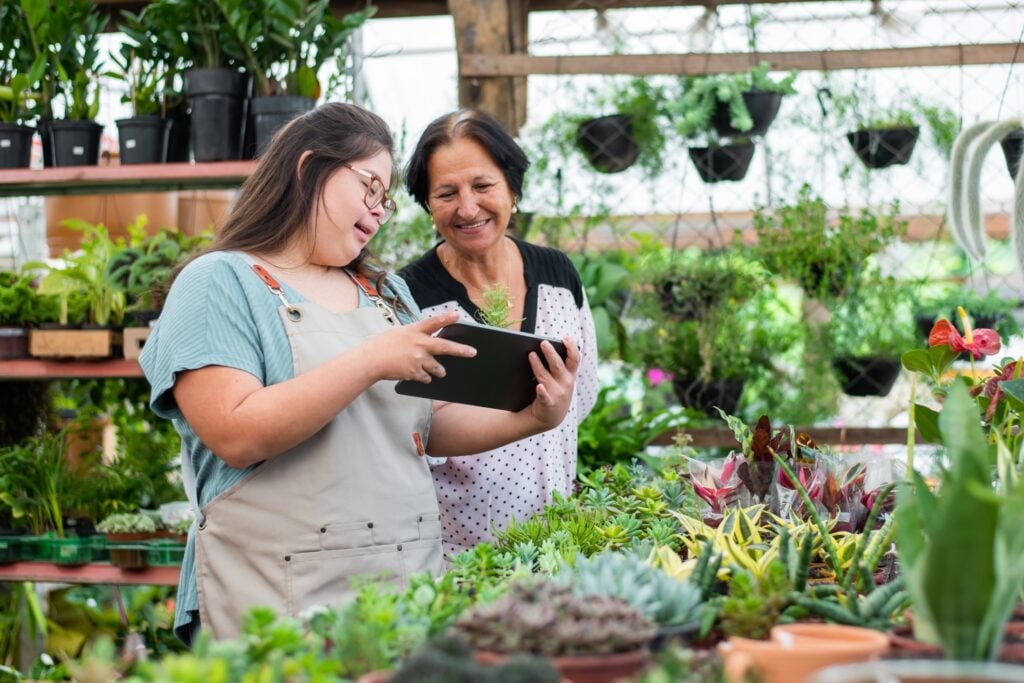 A young woman wearing glasses and a light blue cardigan over an apron works alongside an older woman in a polka dot apron in a bright greenhouse filled with lush plants. They are both smiling while looking at a tablet together, surrounded by various potted plants, succulents, and flowers on wooden shelving and tables. The greenhouse has large windows letting in natural light, creating a warm, nurturing environment for plant care and gardening activities.