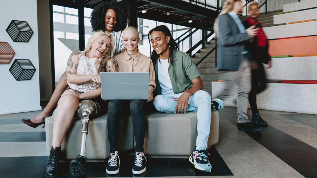 Four diverse people sit closely on a bench in a modern office, smiling and engaged while looking at a laptop, conveying collaboration and positivity.