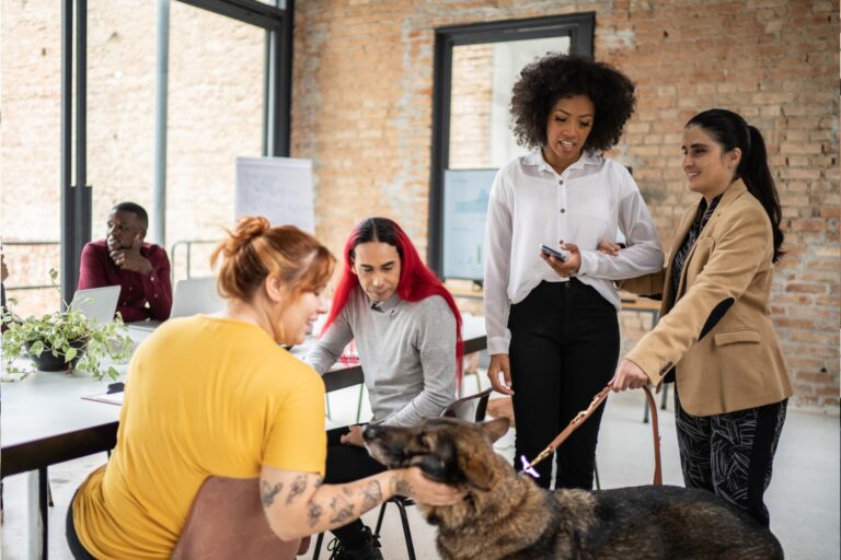 A group of diverse professionals gather around a large dog in a modern office space with exposed brick walls and large windows. A woman in a yellow top with tattoos pets the dog while seated, as colleagues look on with smiles. The group includes a woman with curly hair in a white blouse holding a phone, another woman in a tan blazer, and a woman with red-streaked hair in a gray sweater. In the background, a man in a burgundy shirt works at a laptop. The dog appears to be a German Shepherd or similar breed, creating a pet-friendly workplace atmosphere in this contemporary office setting.