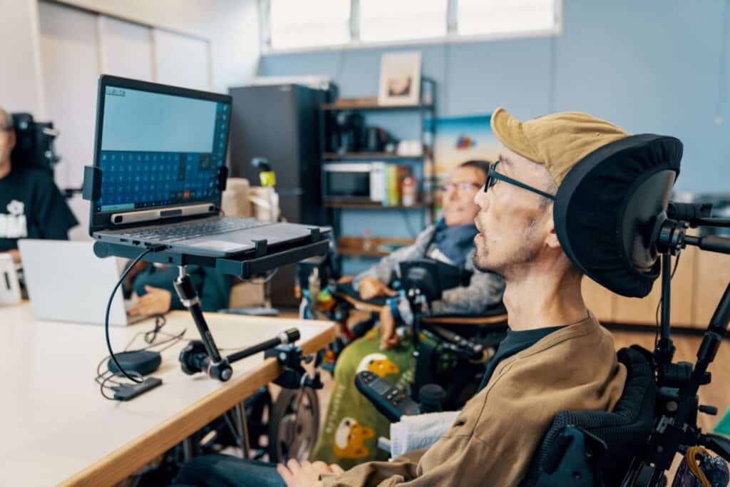 A man with blonde hair and glasses wearing a tan sweater sits in a wheelchair at an accessible workstation, focused on a laptop computer mounted on an adjustable arm. He wears large black headphones and appears to be working or listening to audio content. The modern office space features blue walls, large windows, and shelving with books and supplies. Another person can be seen blurred in the background, creating an inclusive workplace environment designed for accessibility and productivity.