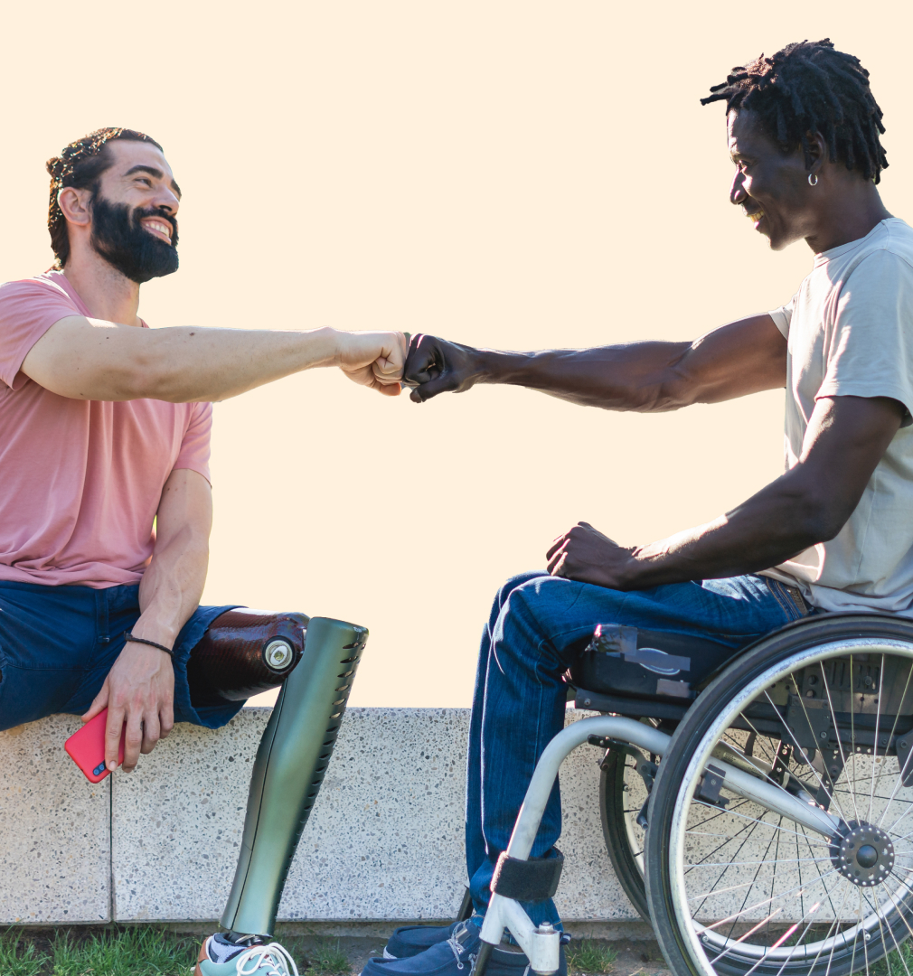 Two men are smiling and fist-bumping; one has a prosthetic leg, and the other is in a wheelchair. They sit outdoors, conveying friendship and positivity.