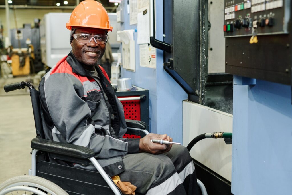 A smiling man in a wheelchair wearing an orange hard hat, safety glasses, and gray work uniform with red accents operates industrial machinery in a manufacturing facility. He holds a clipboard or tablet while positioned at a large blue and gray machine with control panels and various gauges. The industrial setting features overhead lighting and other equipment visible in the background, demonstrating an inclusive workplace where accessibility accommodations allow full participation in skilled manufacturing work.